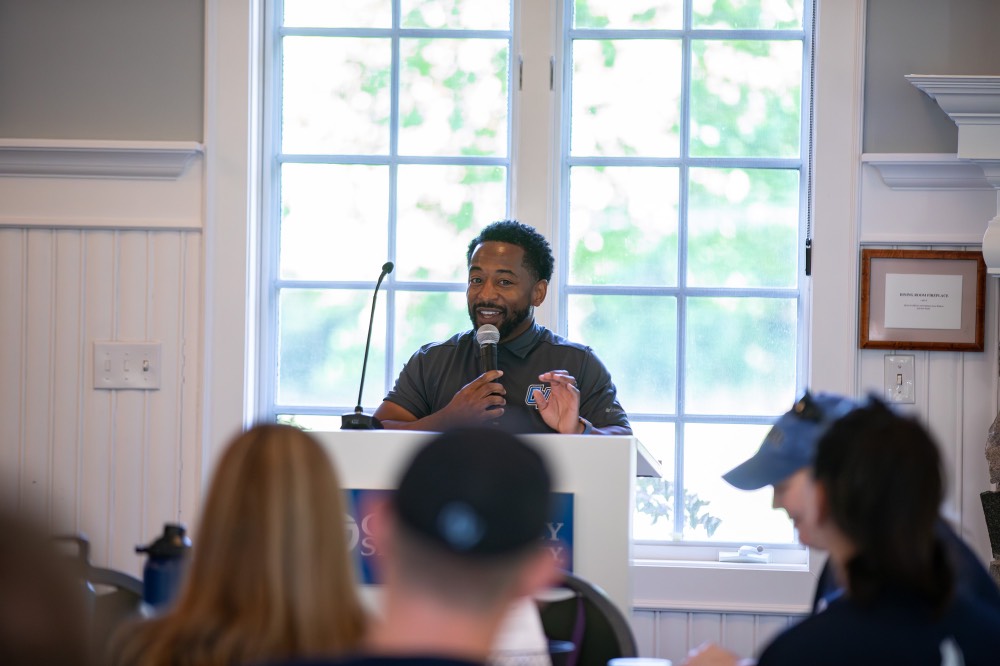 GVSU employee smiles as he holds a microphone talking to the group of GVSU move in day volunteers
